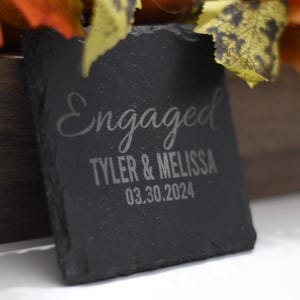 Puede incluir: Un posavasos cuadrado de pizarra negra con la palabra "Engaged" en escritura elegante. Debajo, los nombres "Tyler & Melissa" y la fecha "03.30.2024" están grabados. Un regalo de boda o compromiso.