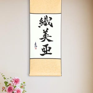 Puede incluir: Un pergamino vertical con caligrafía japonesa en tinta negra. El texto está centrado sobre un fondo blanco, enmarcado por un borde beige. Se ve un pequeño sello rojo. El pergamino cuelga de una pared clara, con un jarrón de flores rosas en primer plano.