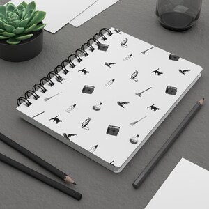 Puede incluir: Un cuaderno blanco con espiral con un patrón negro repetido de iconos temáticos de brujas, incluyendo gatos, sombreros y botellas de pociones. El cuaderno está sobre una superficie gris con lápices y una pequeña planta suculenta en maceta.