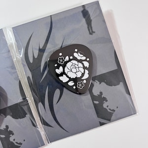 以下が含まれることがあります： 白い花のデザインが施された黒いギターピック。ピックは、黒と白のデザインが施されたグレーの背景の上に置かれています。