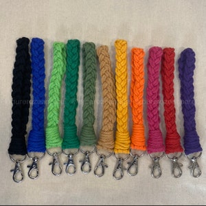 May include: A collection of braided rope keychains in various colors, including black, blue, green, and orange. Each keychain has a silver-colored clasp. The keychains are displayed against a neutral background.