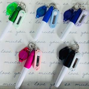 May include: Six white marker pens with different colored tops and attached keychains. Each keychain has a small utility knife and a small light. The tops are green, blue, pink, black, purple, and blue.