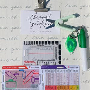 May include: A white card with the text "beyond grateful" and "THANK YOU SO MUCH" written in black ink. The card is clipped to a green plastic tool. A green pen, a green keychain with a small green circle, and two white cards with ECG charts are also visible.