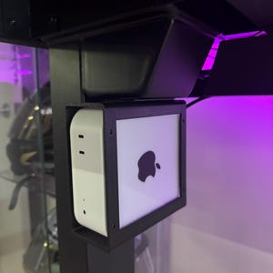 May include: A white Apple TV device with a black frame is mounted to a black desk. The Apple logo is visible on the front of the device.