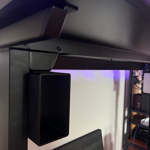 Puede incluir: Escritorio de metal negro con un sistema de gestión de cables negro y un contenedor de almacenamiento negro fijado a la parte inferior del escritorio. Un dispositivo blanco con un logotipo de Apple está fijado a la pata del escritorio.