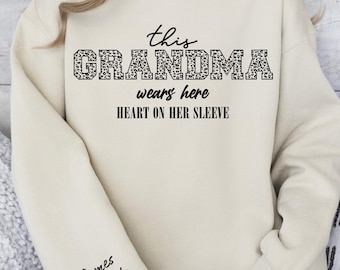 May include: Sand-colored sweatshirt with the text "this GRANDMA wears here HEART ON HER SLEEVE." The word "GRANDMA" is in a leopard print pattern. The sleeve has the name "James Miranda" and a heart.