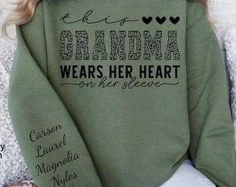 May include: Military green sweatshirt with the text "this GRANDMA wears her heart on her sleeve." The word "GRANDMA" is in a leopard print pattern. The left sleeve has the names "Carson, Laurel, Magnolia, Nyles" in a script font.