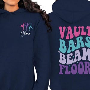 May include: Navy blue hoodie with gymnastics-themed graphics. The front features a gymnast silhouette and the name "Clara". The back displays the words "VAULT BARS BEAM FLOOR" in a retro font, with stars.