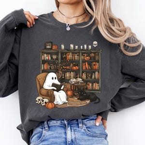May include: Dark gray long-sleeve sweatshirt featuring a Halloween-themed graphic. The graphic depicts a ghost reading in a cozy armchair in front of a bookshelf, with pumpkins, a black cat, and skulls. The sweatshirt is made of a soft material.