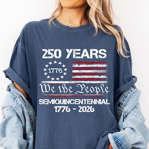 May include: A denim blue t-shirt with the text "250 YEARS" above a distressed American flag graphic. Below the flag, the text reads "We the People" and "Semiquincentennial 1776-2026". The shirt has rolled-up sleeves.