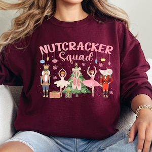 May include: Maroon sweatshirt with the text "Nutcracker Squad" in pink. The graphic features a nutcracker, a ballerina, a Christmas tree, and other festive elements. The sweatshirt is a crew neck style.
