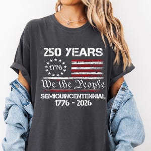 May include: Dark gray t-shirt with a distressed American flag design. The shirt features the text "250 YEARS" above the flag, and "We the People" below. The shirt also includes the text "SEMIQUINCENTENNIAL 1776-2026".