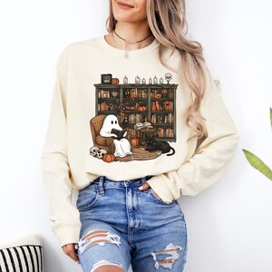 May include: Cream-colored long-sleeve sweatshirt featuring a Halloween-themed graphic. The graphic depicts a ghost reading in a cozy armchair in front of a bookshelf, with pumpkins, candles, and a black cat. The design has a whimsical, cartoonish style.