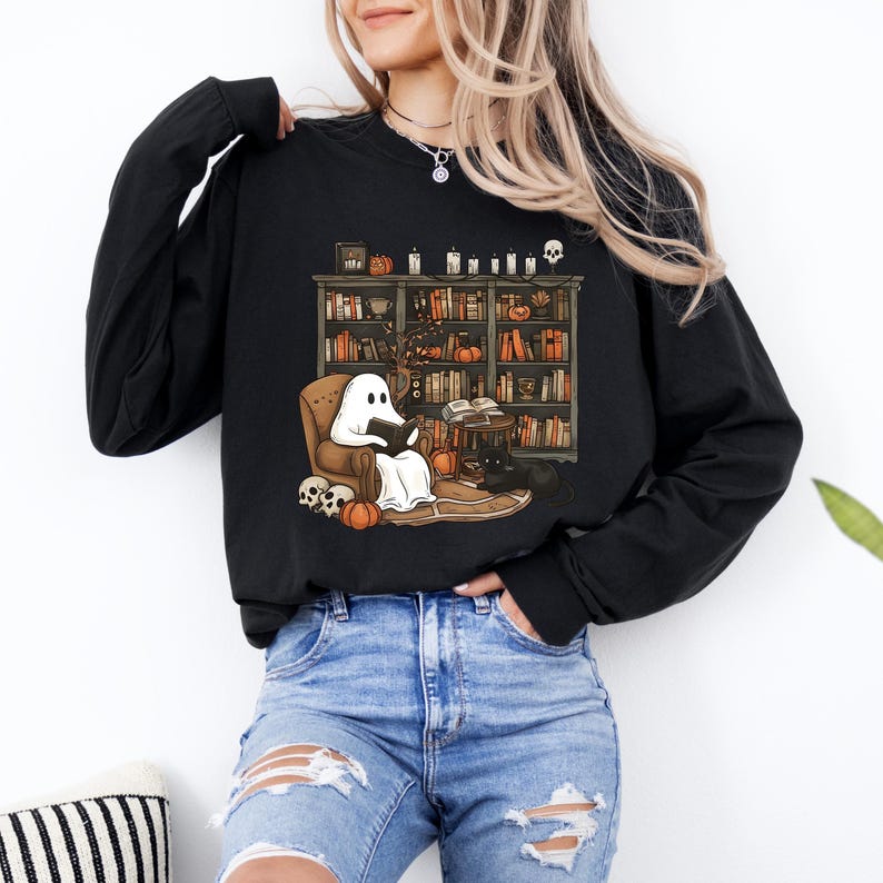 May include: Black long-sleeve shirt featuring a Halloween-themed graphic. The graphic depicts a ghost reading in a cozy chair in front of a bookshelf, with pumpkins, skulls, and a black cat. The design has a whimsical, cartoonish style.