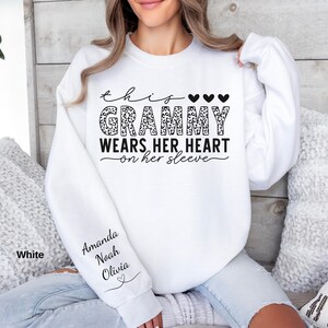 May include: White sweatshirt with the text "This Grammy Wears Her Heart on Her Sleeve" in black script and leopard print. The names Amanda, Noah, and Olivia are printed on the sleeve. Three black hearts are above the word "Grammy".