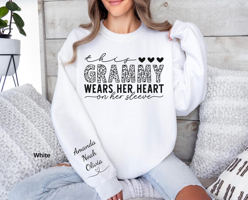May include: White sweatshirt with the text "This Grammy Wears Her Heart on Her Sleeve" in black script and leopard print. The names Amanda, Noah, and Olivia are printed on the sleeve. Three black hearts are above the word "Grammy".