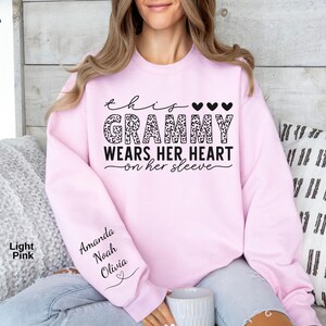 May include: Light pink sweatshirt with the text "This Grammy Wears Her Heart on Her Sleeve." The word "Grammy" is in a leopard print design. The sleeve has the names "Amanda, Noah, Olivia" printed on it.
