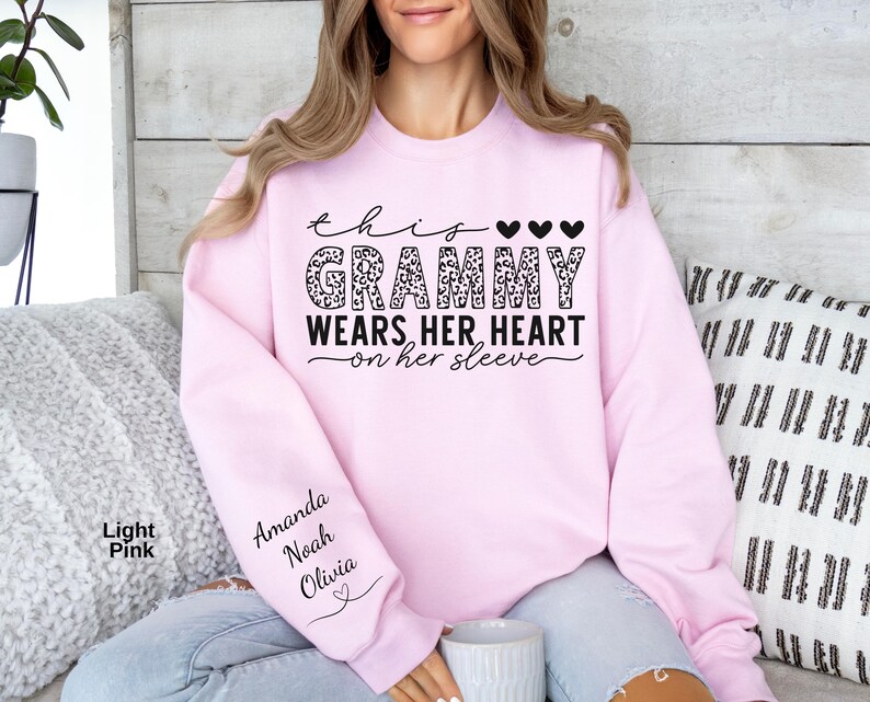 May include: Light pink sweatshirt with the text "This Grammy Wears Her Heart on Her Sleeve." The word "Grammy" is in a leopard print design. The sleeve has the names "Amanda, Noah, Olivia" printed on it.