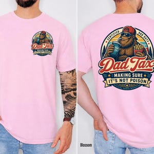 Puede incluir: Camiseta rosa claro con un gr&aacute;fico en la parte delantera y trasera. El gr&aacute;fico frontal es un dise&ntilde;o circular con el texto "Dad Tax" y un Bigfoot de dibujos animados. El gr&aacute;fico trasero es una versi&oacute;n m&aacute;s grande del mismo dise&ntilde;o, con el texto "MAKING SURE IT'S NOT POISON".