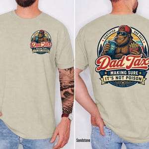 Puede incluir: Camiseta color arena con un gr&aacute;fico de Bigfoot y el texto "Dad Tax" y "Making Sure It's Not Poison". El dise&ntilde;o incluye un Bigfoot sosteniendo una bebida y papas fritas. La camiseta tiene cuello redondo y mangas cortas.