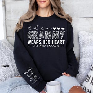 May include: Black sweatshirt with the text "this Grammy wears her heart on her sleeve." The word "Grammy" is in a leopard print design. The names "Amanda, Noah, Olivia" are printed on the left sleeve. The sweatshirt is a crew neck.