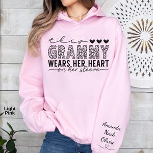May include: Light pink hoodie with the text "This Grammy Wears Her Heart on Her Sleeve." The word "Grammy" is in a leopard print design. The names "Amanda, Noah, Olivia" are printed on the sleeve.