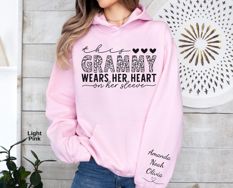 May include: Light pink hoodie with the text "This Grammy Wears Her Heart on Her Sleeve." The word "Grammy" is in a leopard print design. The names "Amanda, Noah, Olivia" are printed on the sleeve.