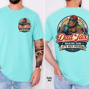 Puede incluir: Camiseta turquesa clara con un gr&aacute;fico de Bigfoot que dice "Dad Tax" y "Making Sure It's Not Poison". El gr&aacute;fico est&aacute; en la parte delantera y trasera de la camiseta. El gr&aacute;fico trasero tambi&eacute;n dice "Certified Bad Duty" y "Official Snack Inspector."