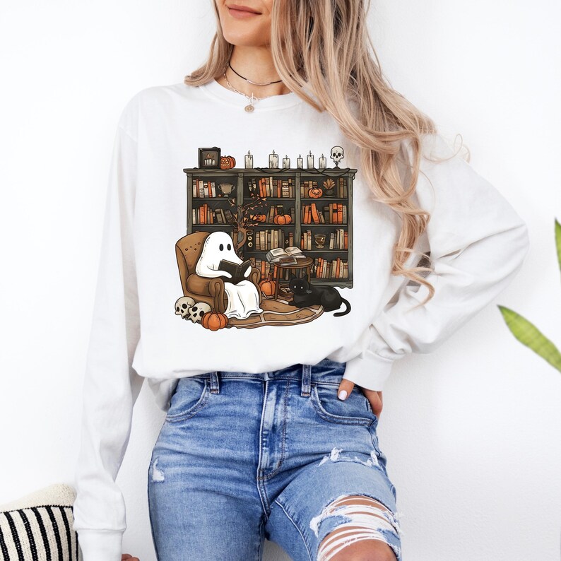 May include: White long-sleeve shirt featuring a Halloween-themed illustration. The design shows a ghost reading in a cozy chair in front of a bookshelf, with pumpkins, a black cat, and skulls. A fun and festive design for the spooky season.
