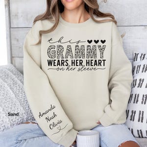 May include: Sand-colored sweatshirt with the text "This Grammy Wears Her Heart on Her Sleeve." The word "Grammy" has a leopard print design. The sleeve has the names "Amanda, Noah, Olivia" printed on it.