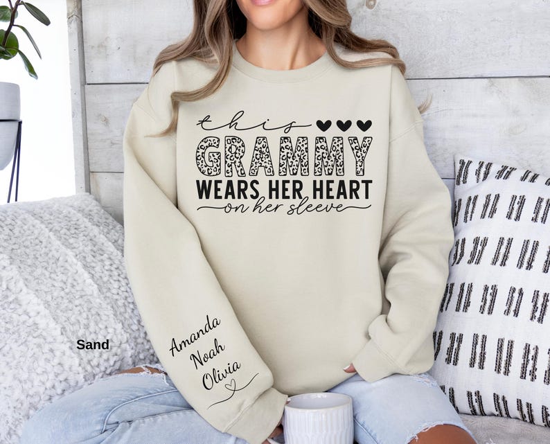 May include: Sand-colored sweatshirt with the text "This Grammy Wears Her Heart on Her Sleeve." The word "Grammy" has a leopard print design. The sleeve has the names "Amanda, Noah, Olivia" printed on it.