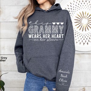 May include: Dark grey heather hoodie with the text "this Grammy wears her heart on her sleeve" in white script. The word "Grammy" has a leopard print design. The names Amanda, Noah, and Olivia are printed on the sleeve.