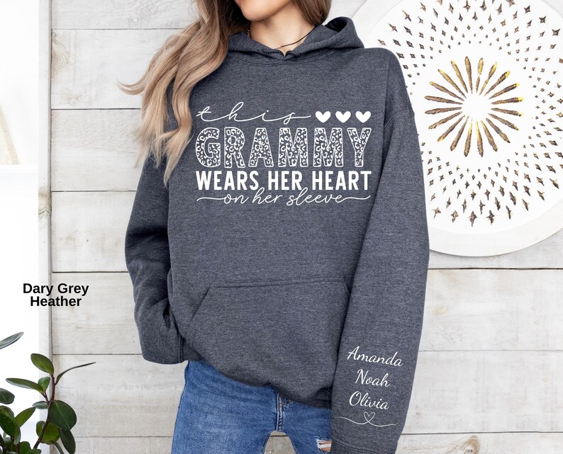 May include: Dark grey heather hoodie with the text "this Grammy wears her heart on her sleeve" in white script. The word "Grammy" has a leopard print design. The names Amanda, Noah, and Olivia are printed on the sleeve.
