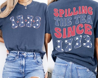 May include: A navy blue t-shirt with the number "1773" on the front, filled with a white star pattern and red outlines. The back of the shirt says "Spilling the Tea Since 1773" in red and blue bubble letters, with a star-filled "1773" below.
