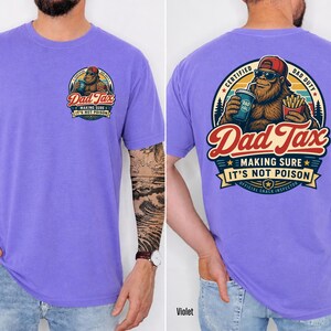 Puede incluir: Camiseta violeta de manga corta con un gr&aacute;fico en la parte delantera y trasera. El gr&aacute;fico presenta un personaje parecido a Bigfoot con gafas de sol y un sombrero rojo, sosteniendo una bebida y papas fritas. El texto incluye "Dad Tax" y "Making Sure It's Not Poison."