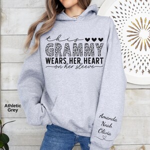 May include: Athletic grey hoodie with the text "This Grammy Wears Her Heart on Her Sleeve." The word "Grammy" is in a leopard print design. The sleeve has the names "Amanda, Noah, Olivia" and a heart.