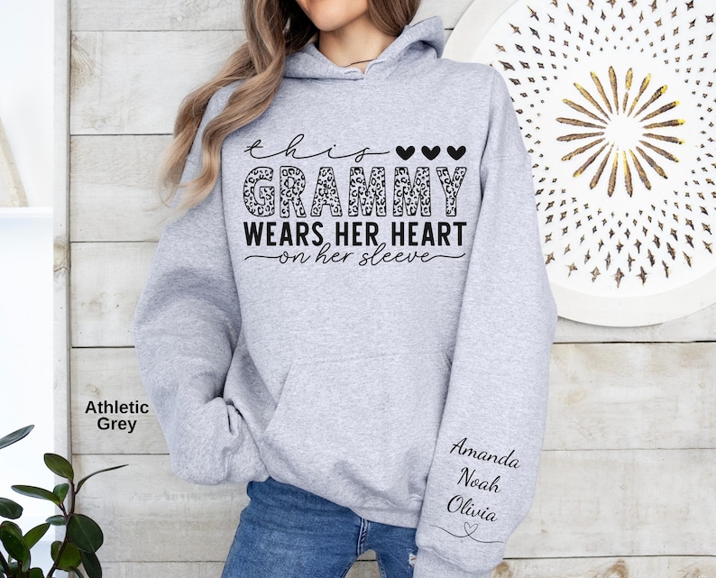 May include: Athletic grey hoodie with the text "This Grammy Wears Her Heart on Her Sleeve." The word "Grammy" is in a leopard print design. The sleeve has the names "Amanda, Noah, Olivia" and a heart.