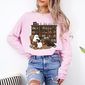 May include: A light pink long-sleeve sweatshirt featuring a Halloween-themed graphic. The graphic depicts a ghost reading in a cozy chair in front of a bookshelf, with pumpkins, skulls, and a black cat. The sweatshirt is paired with blue jeans.