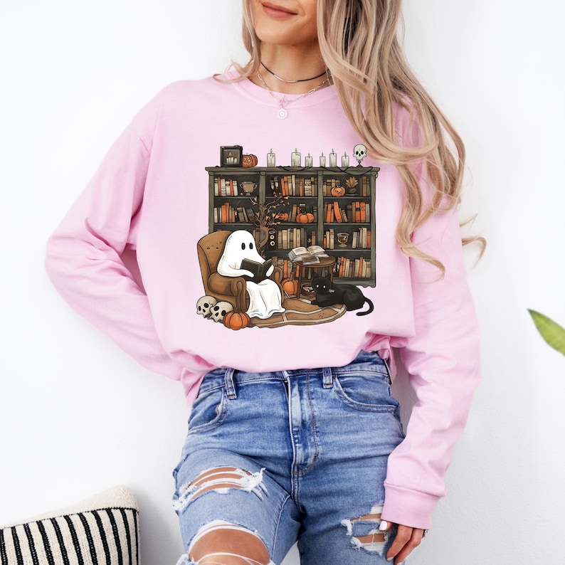 May include: A light pink long-sleeve sweatshirt featuring a Halloween-themed graphic. The graphic depicts a ghost reading in a cozy chair in front of a bookshelf, with pumpkins, skulls, and a black cat. The sweatshirt is paired with blue jeans.