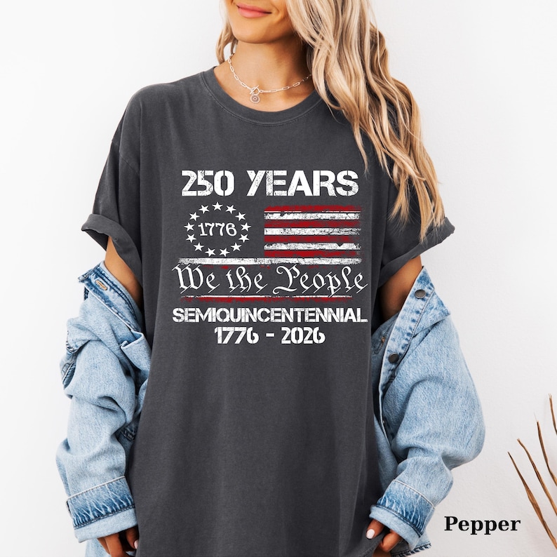 May include: Dark gray t-shirt with a distressed American flag design. The shirt features the text "250 YEARS" above the flag, and "We the People" below. The shirt also includes the text "SEMIQUINCENTENNIAL 1776-2026".