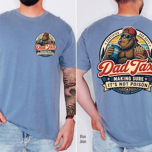 Puede incluir: Camiseta azul claro con un dise&ntilde;o de dibujos animados de Bigfoot. La parte delantera muestra un peque&ntilde;o gr&aacute;fico con el texto "Dad Tax". La parte trasera muestra un gr&aacute;fico m&aacute;s grande de Bigfoot con las palabras "Dad Tax, Making Sure It's Not Poison".