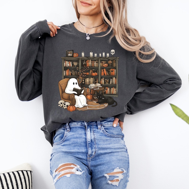 May include: Dark gray long-sleeve sweatshirt featuring a Halloween-themed graphic. The graphic depicts a ghost reading in a cozy armchair in front of a bookshelf, with pumpkins, a black cat, and skulls. The sweatshirt is made of a soft material.