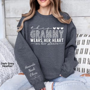 May include: Dark gray heather sweatshirt with the text "this grammy wears her heart on her sleeve" in white. The word "grammy" is in a leopard print pattern. The names Amanda, Noah, and Olivia are printed on the sleeve.