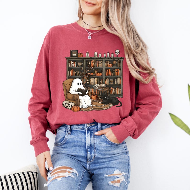 May include: A maroon long-sleeve shirt featuring a graphic of a ghost reading in a cozy library setting. The design includes a bookshelf, pumpkins, a black cat, and skulls, creating a Halloween or autumn theme. The shirt is paired with blue jeans.