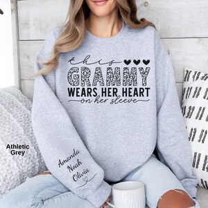 May include: Athletic grey sweatshirt with the text "This Grammy Wears Her Heart on Her Sleeve." The word "Grammy" has a leopard print design. The sleeve has the names Amanda, Noah, and Olivia.