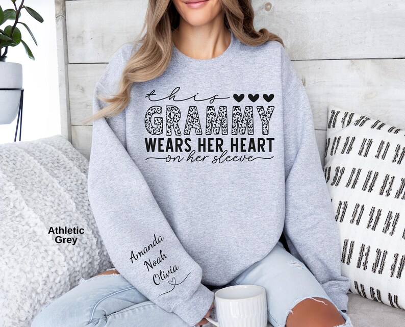 May include: Athletic grey sweatshirt with the text "This Grammy Wears Her Heart on Her Sleeve." The word "Grammy" has a leopard print design. The sleeve has the names Amanda, Noah, and Olivia.