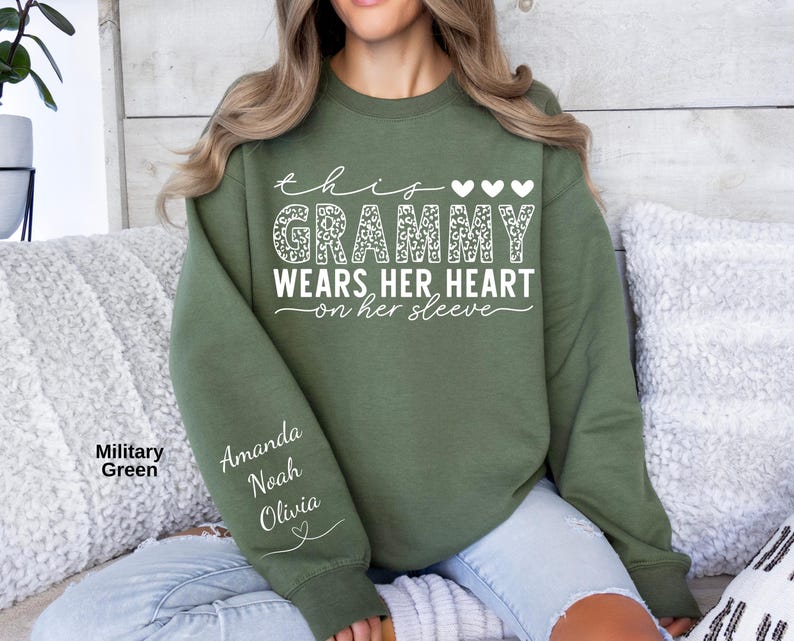 May include: A military green sweatshirt with the text "this Grammy wears her heart on her sleeve" in white. The word "Grammy" has a leopard print design. The sleeve has the names "Amanda, Noah, Olivia" in white.