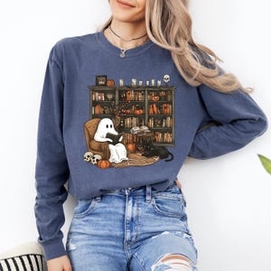 May include: A blue sweatshirt featuring a whimsical illustration of a ghost reading in a cozy armchair in front of a bookshelf. The design includes pumpkins, a black cat, and skulls, perfect for Halloween or book lovers.