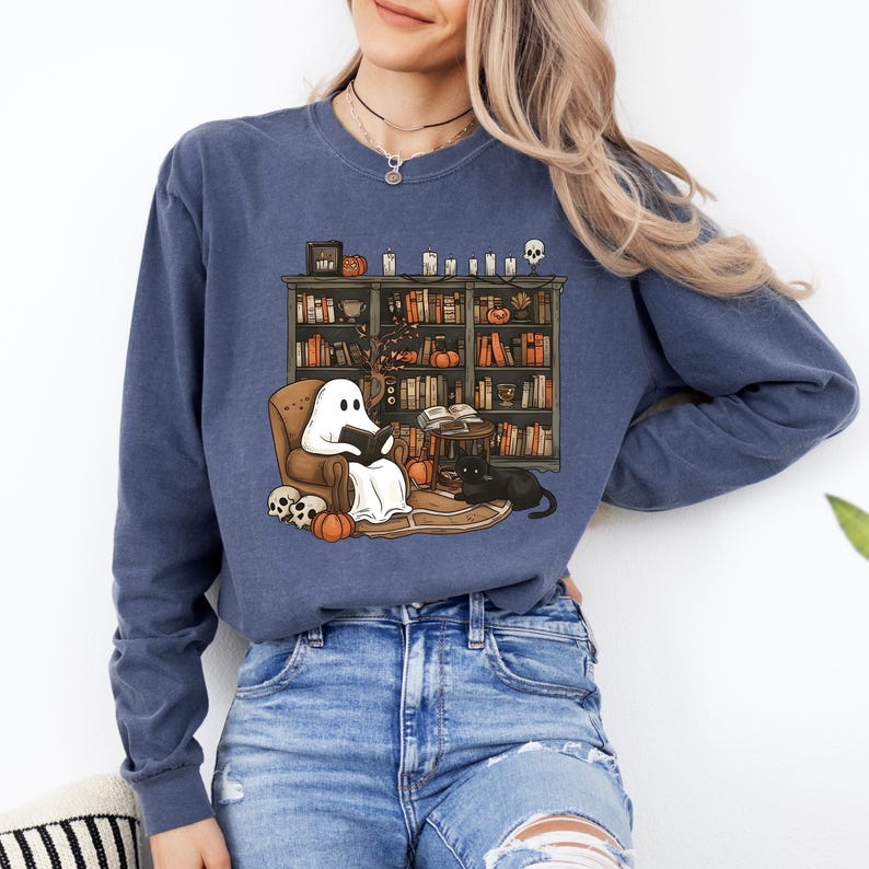 May include: A blue sweatshirt featuring a whimsical illustration of a ghost reading in a cozy armchair in front of a bookshelf. The design includes pumpkins, a black cat, and skulls, perfect for Halloween or book lovers.