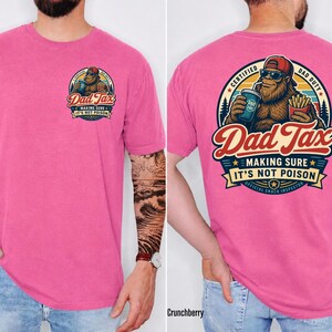 Puede incluir: Una camiseta rosa con un gr&aacute;fico en la parte delantera y trasera. El gr&aacute;fico frontal dice "Dad Tax" con un personaje tipo Pie Grande. El gr&aacute;fico trasero presenta el mismo personaje, el texto "Dad Tax" y la frase "Making Sure It's Not Poison."